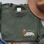 Military Green crewneck sweatshirt laid flat with a felt hat, embroidered on the left chest with a long-haired dachshund in a ghost sheet carrying a pumpkin bucket under the word spooky