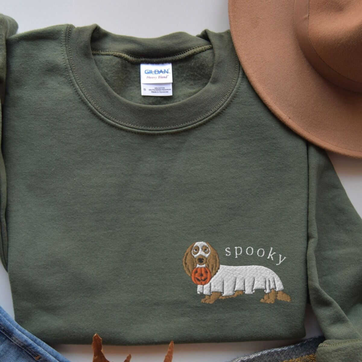 Spooky Dachshund Sweatshirt – Military Green Military Green crewneck sweatshirt laid flat with a felt hat, embroidered on the left chest with a long-haired dachshund in a ghost sheet carrying a pumpkin bucket under the word spooky