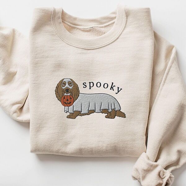 Sand crewneck sweatshirt laid flat on white, embroidered in the center with a long-haired dachshund in a ghost sheet carrying a jack-o'-lantern bucket under the word spooky