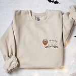 Sand crewneck sweatshirt laid flat among bats and fairy lights, embroidered small on the left chest with a long-haired dachshund in a ghost sheet and the word spooky