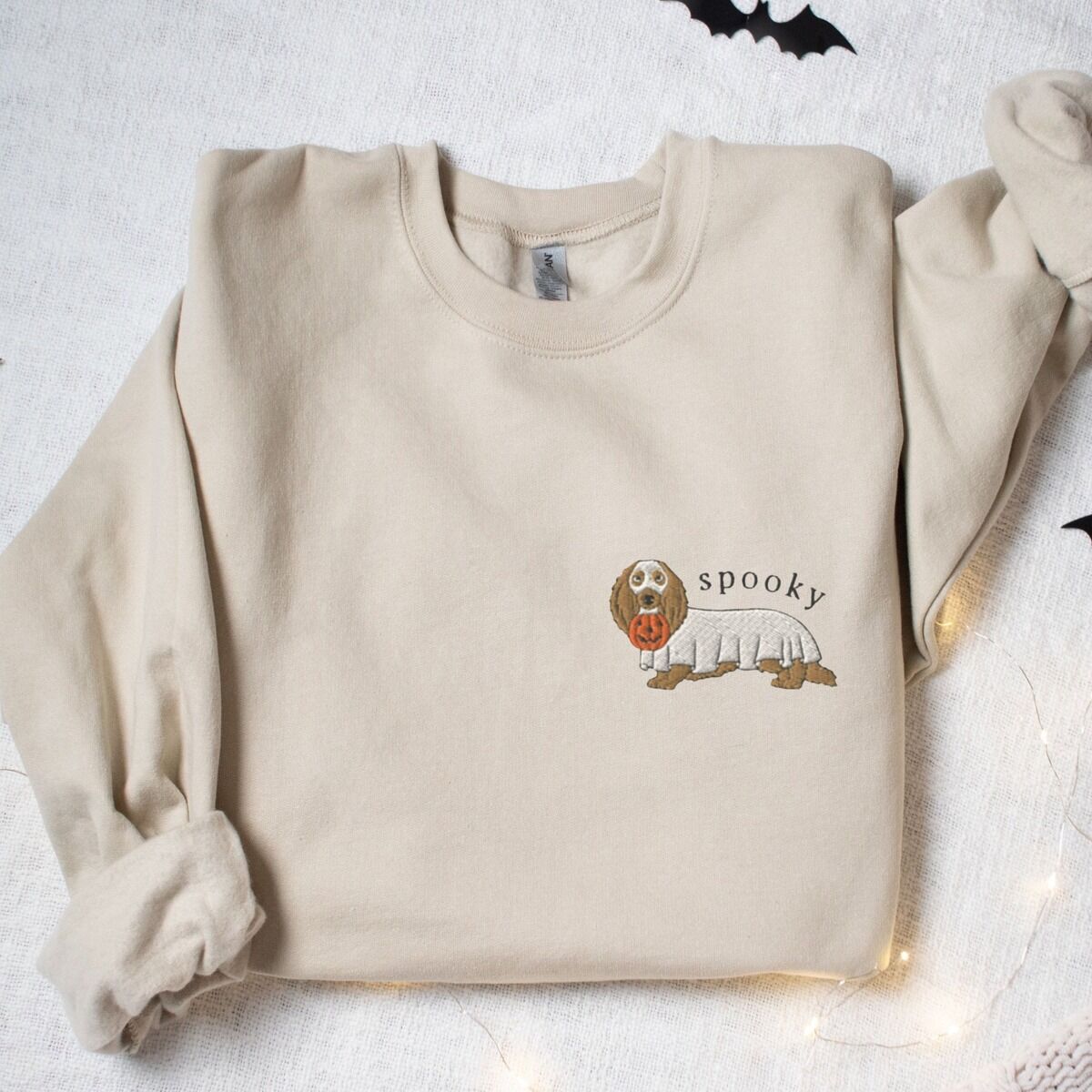 Spooky Dachshund Sweatshirt - left chest on sand Sand crewneck sweatshirt laid flat among bats and fairy lights, embroidered small on the left chest with a long-haired dachshund in a ghost sheet and the word spooky