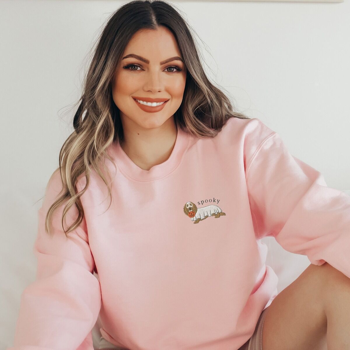 Spooky Dachshund Sweatshirt - left chest worn Woman in a Light Pink crewneck sweatshirt embroidered small on the left chest with a long-haired dachshund in a ghost sheet and the word spooky