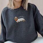Woman in a Dark Heather crewneck sweatshirt embroidered in the center with a long-haired dachshund in a ghost sheet carrying a pumpkin bucket under the word spooky