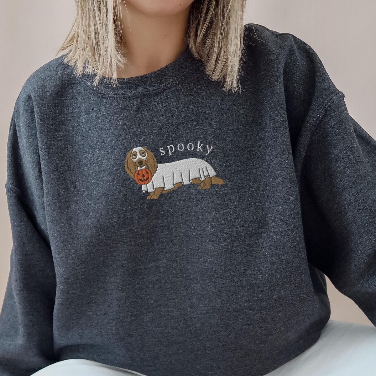 Spooky Dachshund Sweatshirt - worn Woman in a Dark Heather crewneck sweatshirt embroidered in the center with a long-haired dachshund in a ghost sheet carrying a pumpkin bucket under the word spooky