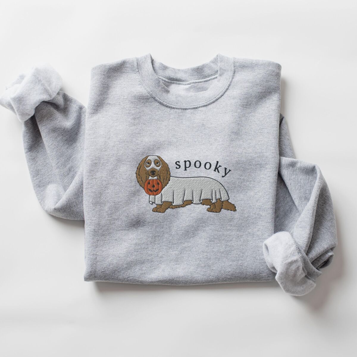 Spooky Dachshund Sweatshirt - Sport Grey Sport Grey crewneck sweatshirt laid flat on white, embroidered in the center with a long-haired dachshund in a ghost sheet carrying a jack-o'-lantern bucket under the word spooky