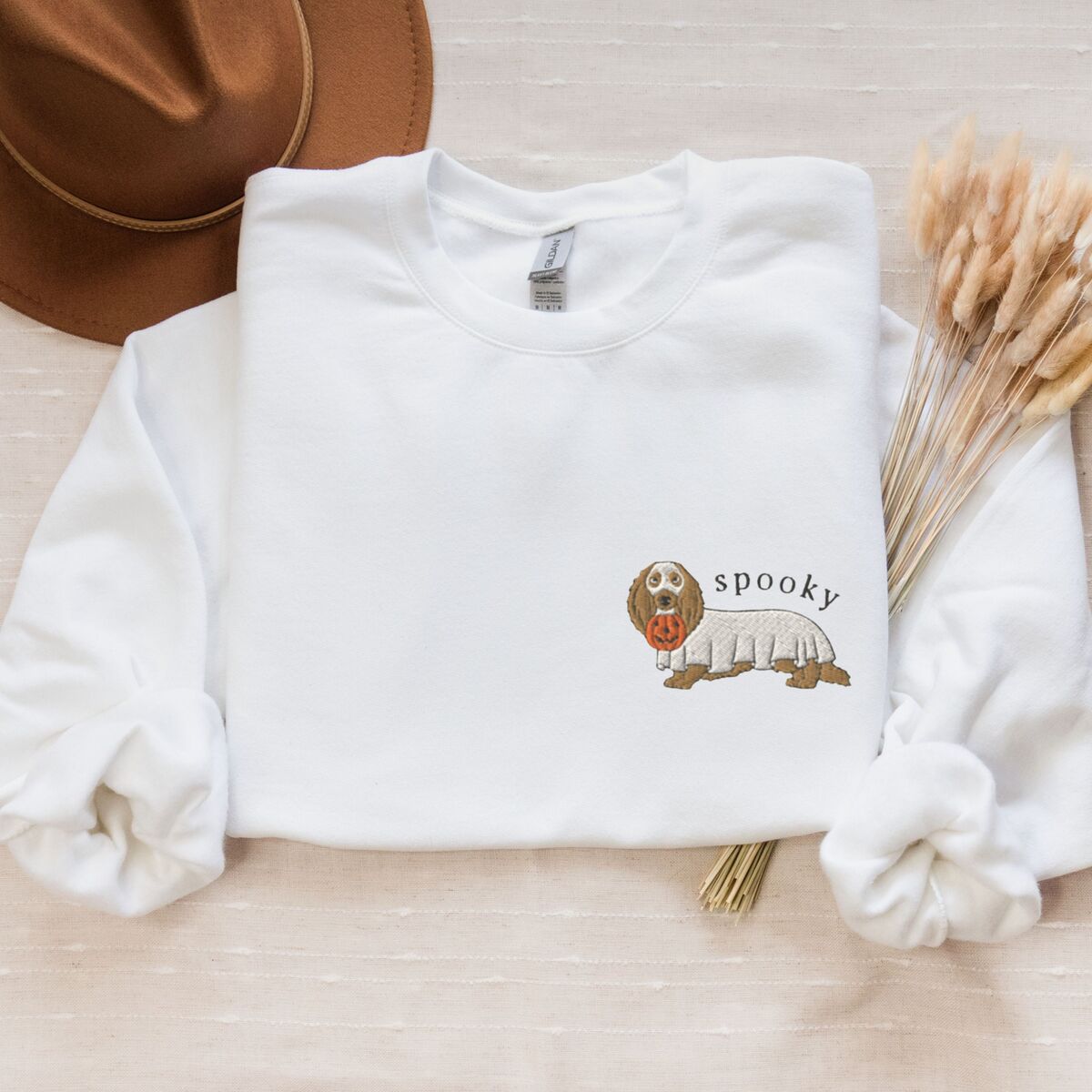 Spooky Dachshund Sweatshirt - White White crewneck sweatshirt laid flat with a felt hat and dried wheat, embroidered on the left chest with a long-haired dachshund in a ghost sheet and the word spooky