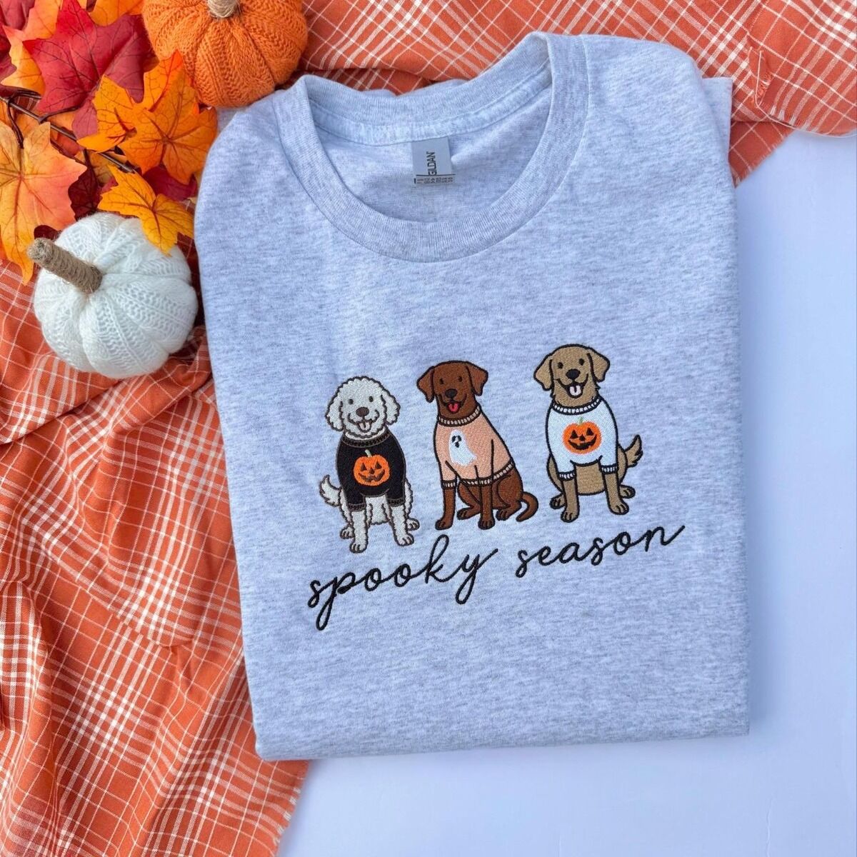 Spooky Pups Sweatshirt – Ash Ash Gildan crewneck sweatshirt folded on an orange plaid blanket with knit pumpkins and fall leaves, embroidered with a doodle, a chocolate lab and a golden retriever in Halloween sweaters above spooky season in black script