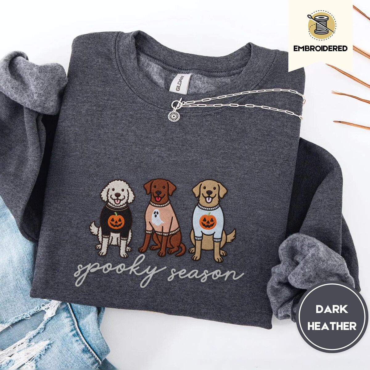 Spooky Pups Sweatshirt – Dark Heather Dark Heather crewneck sweatshirt folded with ripped jeans and a silver chain necklace, embroidered with a doodle, a chocolate lab and a golden retriever in Halloween sweaters above spooky season in script