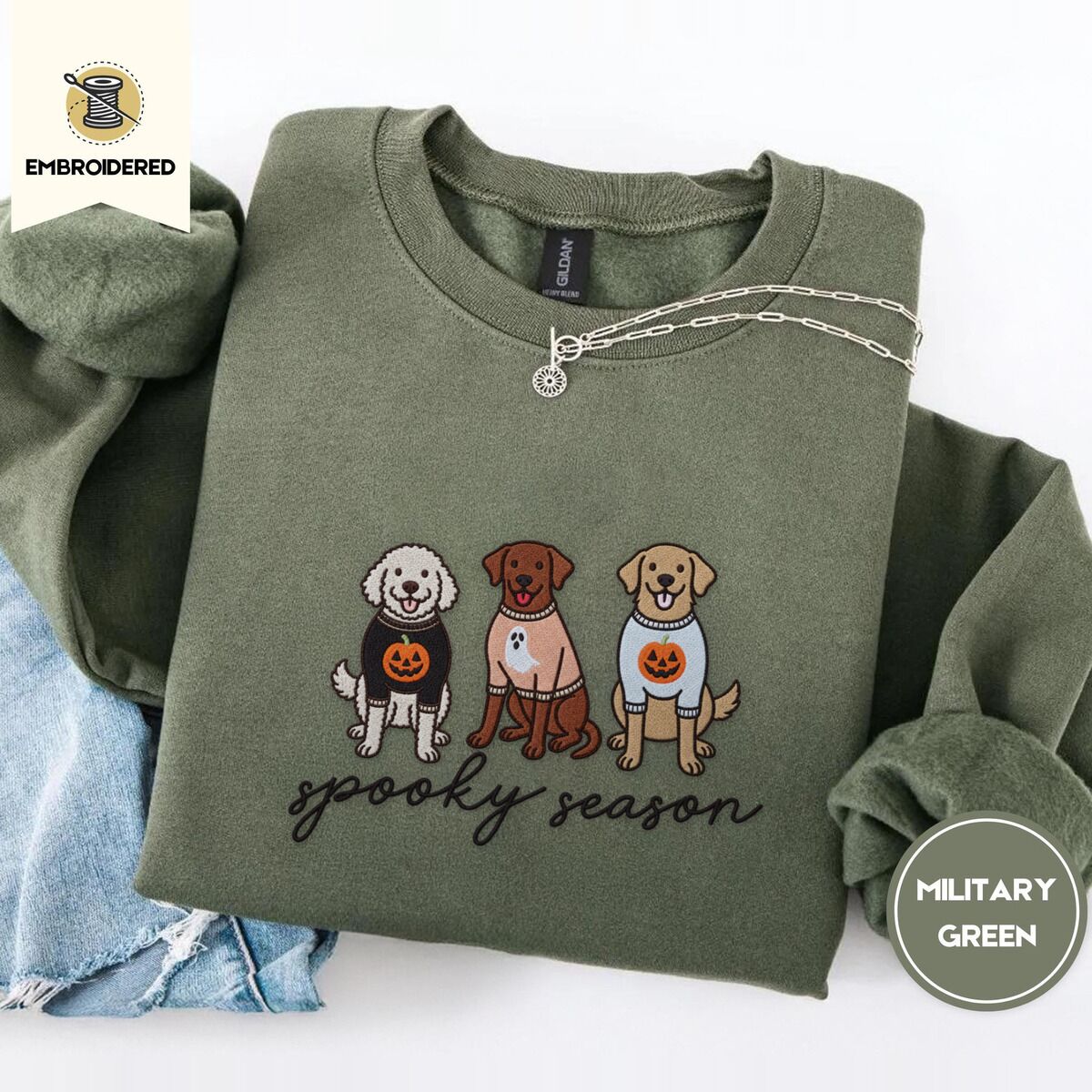 Spooky Pups Sweatshirt – Military Green Military Green crewneck sweatshirt folded with light jeans and a silver chain necklace, embroidered with a doodle, a chocolate lab and a golden retriever in Halloween sweaters above spooky season in script