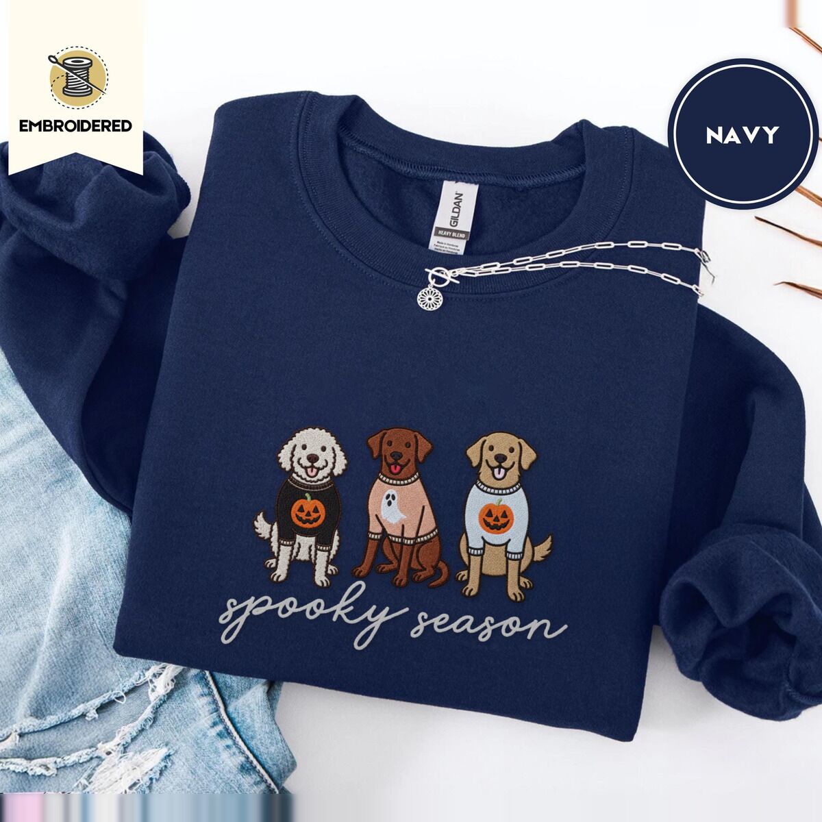 Spooky Pups Sweatshirt – Navy Navy crewneck sweatshirt folded with ripped jeans and a silver chain necklace, embroidered with a doodle, a chocolate lab and a golden retriever in Halloween sweaters above spooky season in script