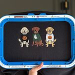 Black sweatshirt held in a blue magnetic embroidery hoop with three freshly stitched dogs in Halloween sweaters, a doodle, a chocolate lab and a golden retriever, shown without the words