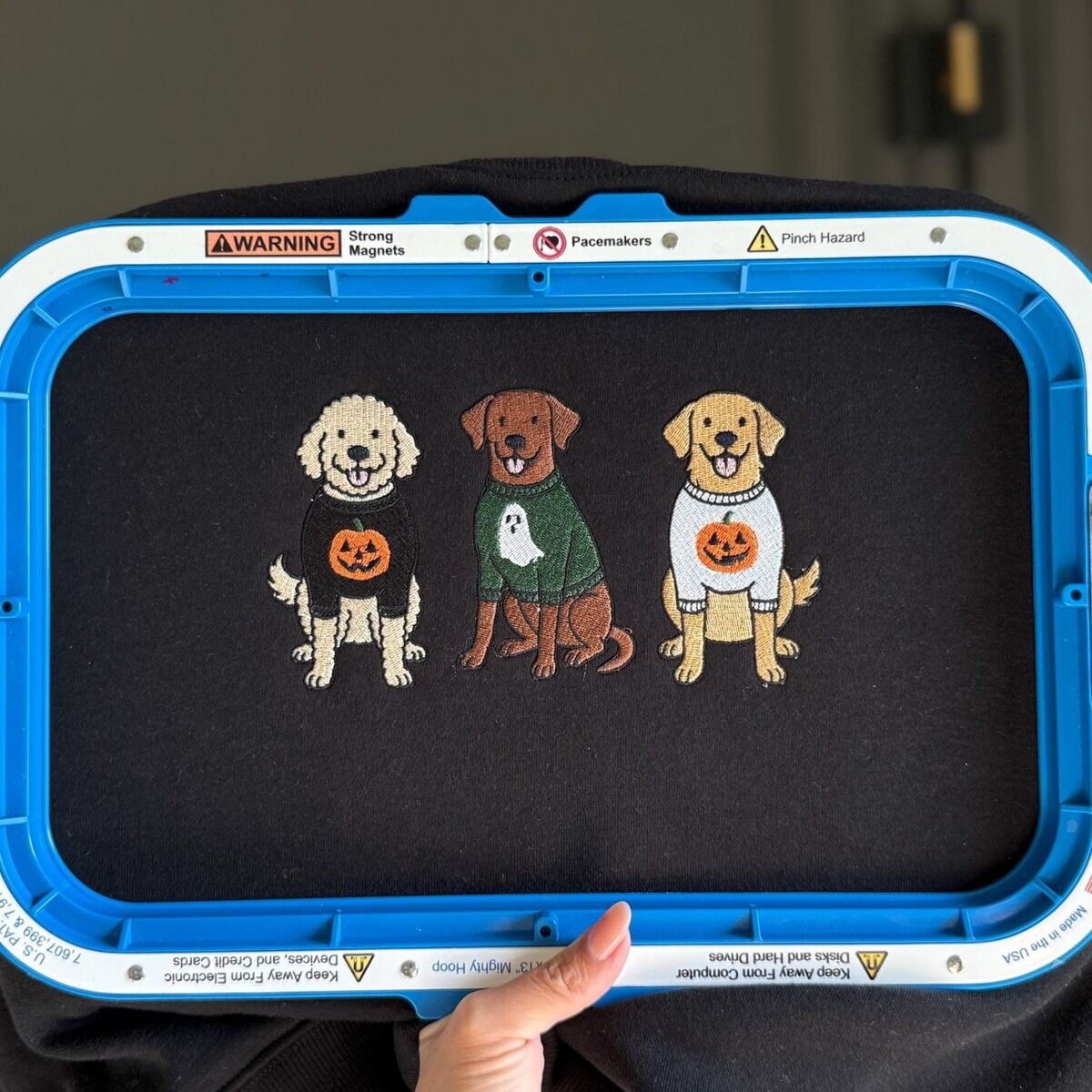 Spooky Pups Embroidery in the Hoop Black sweatshirt held in a blue magnetic embroidery hoop with three freshly stitched dogs in Halloween sweaters, a doodle, a chocolate lab and a golden retriever, shown without the words