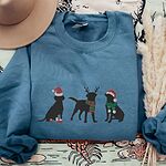 Indigo Blue crewneck sweatshirt laid flat beside a hat and pampas grass, embroidered with three black Labradors in a Santa hat and booties, reindeer antlers with a striped scarf, and a Santa hat with a green scarf