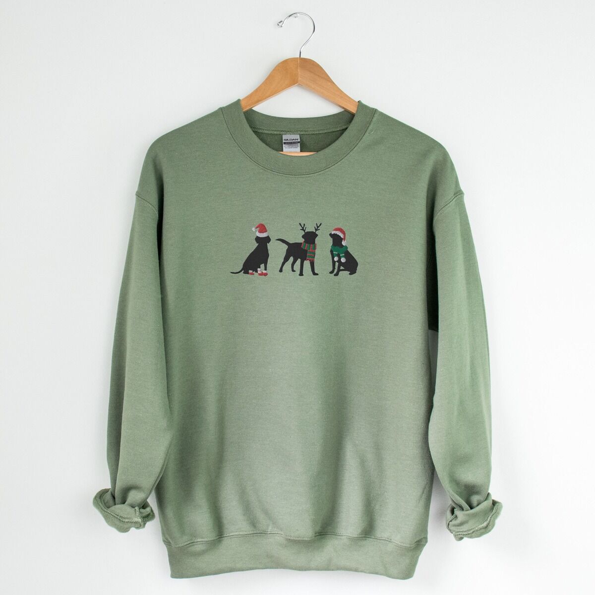 Three Black Labs Christmas Sweatshirt – Military Green Military Green crewneck sweatshirt hanging on a wooden hanger, embroidered with three black Labradors in a Santa hat and booties, reindeer antlers with a striped scarf, and a Santa hat with a green scarf