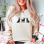 Woman in a cream crewneck sweatshirt in a holiday bedroom, embroidered with three black Labradors in a Santa hat and booties, reindeer antlers with a striped scarf, and a Santa hat with a green scarf