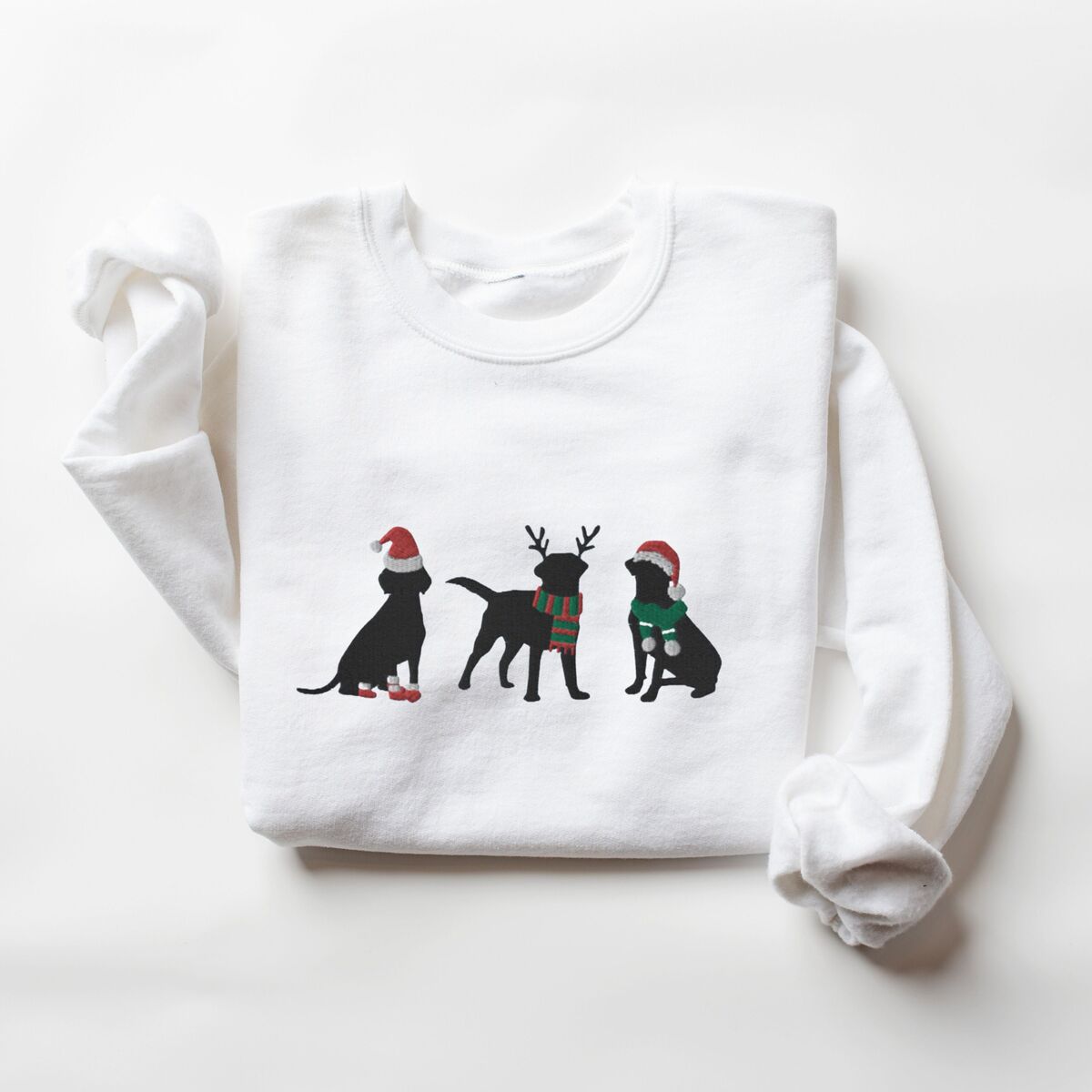 Three Black Labs Christmas Sweatshirt - White White crewneck sweatshirt laid flat with folded sleeves, embroidered with three black Labradors in a Santa hat and booties, reindeer antlers with a striped scarf, and a Santa hat with a green scarf