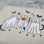 Close-up of three embroidered ghost cats with black tails, one holding an orange jack-o'-lantern, under bats and moons