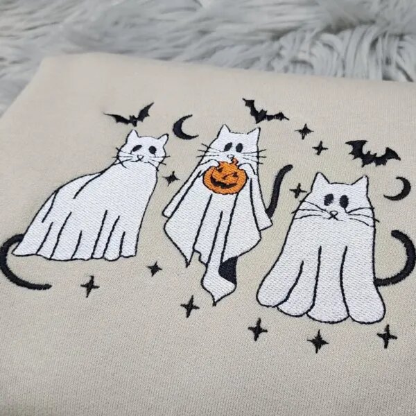 Close-up of three embroidered ghost cats with black tails, one holding an orange jack-o'-lantern, under bats and moons