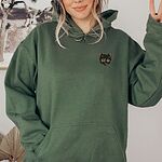 Woman in a Military Green pullover hoodie with one hand in the front pocket, embroidered with a small tortoiseshell cat face, black with golden caramel patches, on the left chest