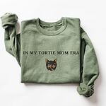 Military Green crewneck sweatshirt laid flat on white with one sleeve bent, embroidered with the words IN MY TORTIE MOM ERA above a tortoiseshell cat face with caramel patches and whiskers, centered on the chest