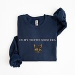 Navy crewneck sweatshirt laid flat on white with one sleeve bent, embroidered with the words IN MY TORTIE MOM ERA in light grey above a tortoiseshell cat face with caramel patches and whiskers, centered on the chest