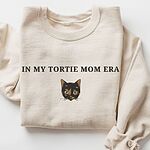 Sand crewneck sweatshirt laid flat with folded sleeves, embroidered with the words IN MY TORTIE MOM ERA above a tortoiseshell cat face with caramel patches and whiskers, centered on the chest
