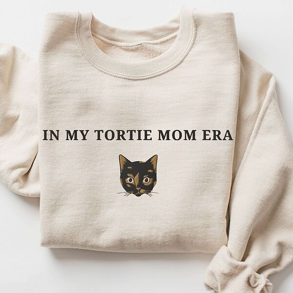 Sand crewneck sweatshirt laid flat with folded sleeves, embroidered with the words IN MY TORTIE MOM ERA above a tortoiseshell cat face with caramel patches and whiskers, centered on the chest