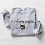 Sport Grey crewneck sweatshirt laid flat with folded sleeves, embroidered with the words IN MY TORTIE MOM ERA above a tortoiseshell cat face with caramel patches and whiskers