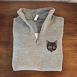 Sport Grey quarter zip pullover folded on a wooden table with the zipper open, embroidered with a small tortoiseshell cat face, black with golden caramel patches, on the left chest