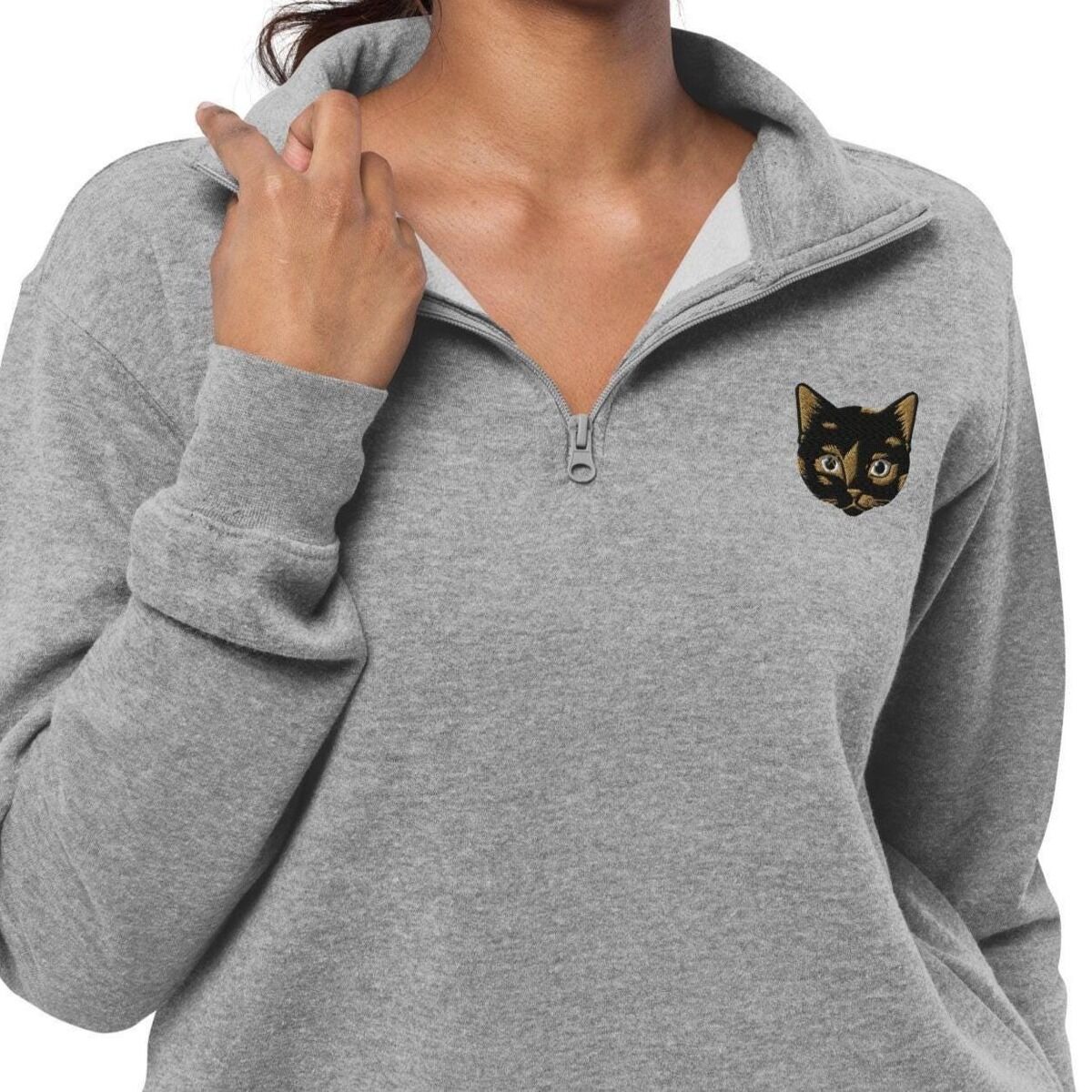 Tortoiseshell Cat Quarter Zip - worn Woman holding the zip collar of a heather grey quarter zip pullover embroidered with a small tortoiseshell cat face, black with golden caramel patches, on the left chest