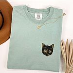Bay T-shirt folded beside a tan hat, a gold necklace and a dried palm leaf, embroidered with a small tortoiseshell cat face, black with golden caramel patches, on the left chest