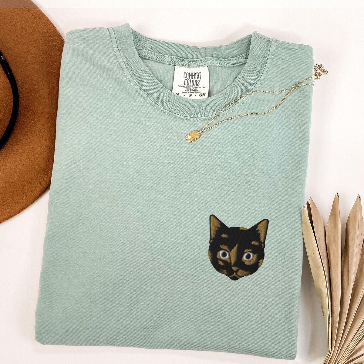Tortoiseshell Cat Shirt – Bay Bay T-shirt folded beside a tan hat, a gold necklace and a dried palm leaf, embroidered with a small tortoiseshell cat face, black with golden caramel patches, on the left chest