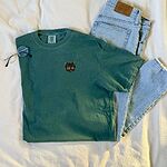 Blue Spruce T-shirt laid on a white blanket next to jeans and glasses, embroidered with a small tortoiseshell cat face, black with golden caramel patches, on the left chest