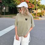 Woman in a white cap and a Khaki T-shirt tucked into white pants on a street, with a small tortoiseshell cat face, black with golden caramel patches, on the left chest
