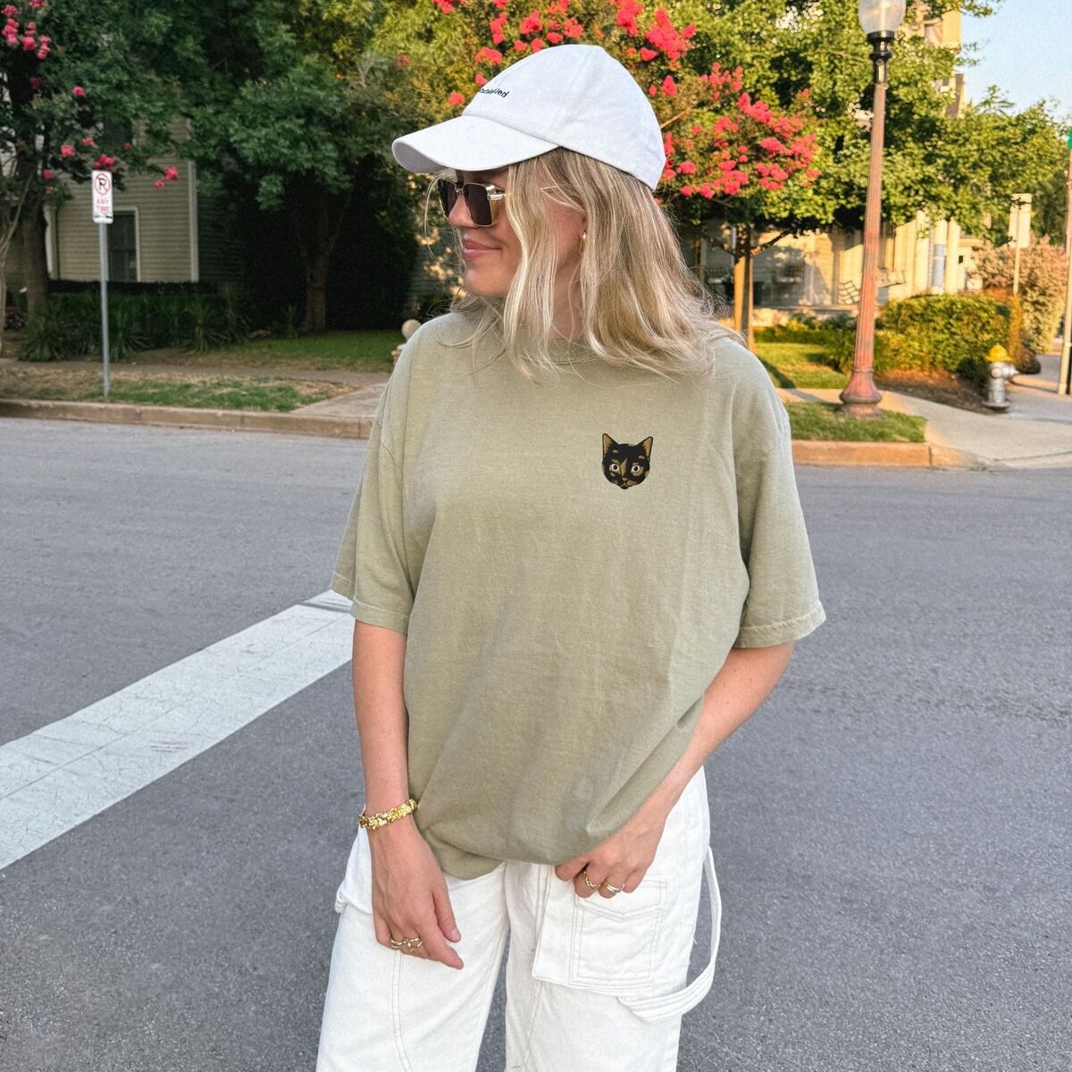 Tortoiseshell Cat Shirt – Khaki Woman in a white cap and a Khaki T-shirt tucked into white pants on a street, with a small tortoiseshell cat face, black with golden caramel patches, on the left chest
