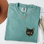 Light Green T-shirt folded beside a tan hat, a gold necklace and a dried palm leaf, embroidered with a small tortoiseshell cat face, black with golden caramel patches, on the left chest