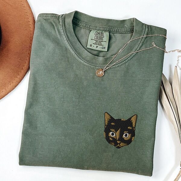 Moss T-shirt folded beside a tan hat, a gold necklace and a dried palm leaf, embroidered with a small tortoiseshell cat face, black with golden caramel patches, on the left chest