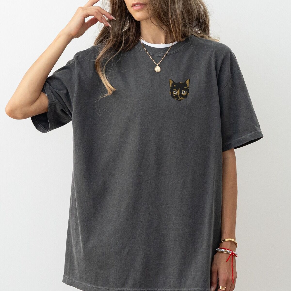 Tortoiseshell Cat Shirt – Pepper Woman in an oversized Pepper T-shirt with one hand in her hair, embroidered with a small tortoiseshell cat face, black with golden caramel patches, on the left chest