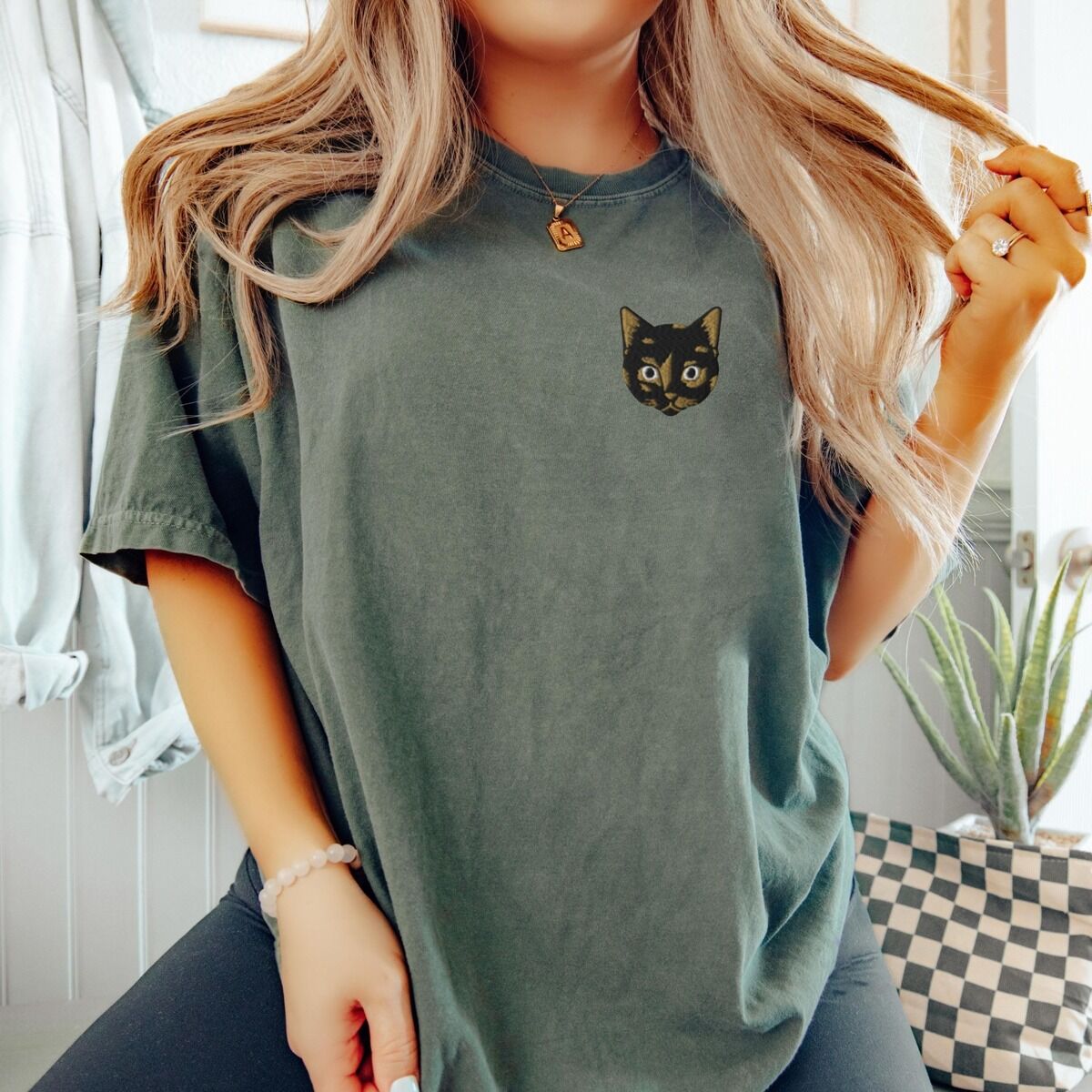 Tortoiseshell Cat Shirt - worn Woman sitting in a Moss T-shirt beside a checkered table, embroidered with a small tortoiseshell cat face, black with golden caramel patches, on the left chest