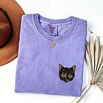 Violet T-shirt folded beside a tan hat, a gold necklace and a dried palm leaf, embroidered with a small tortoiseshell cat face, black with golden caramel patches, on the left chest