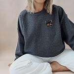 Woman sitting cross-legged in a Dark Heather crewneck sweatshirt and white pants, with a tortoiseshell cat face, black with caramel patches, embroidered on the left chest