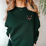 Woman in a Forest Green crewneck sweatshirt and a gold heart necklace, with a tortoiseshell cat face, black with caramel patches, embroidered on the left chest
