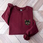 Maroon crewneck sweatshirt folded on a light wood floor with a tortoiseshell cat face, black with caramel patches, embroidered on the left chest