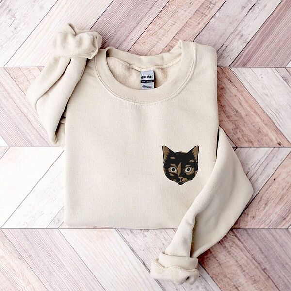 Sand crewneck sweatshirt folded on a chevron wood floor with a tortoiseshell cat face, black with caramel patches, embroidered on the left chest