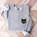 Sport Grey crewneck sweatshirt folded on a chevron wood floor with a tortoiseshell cat face, black with caramel patches, embroidered on the left chest