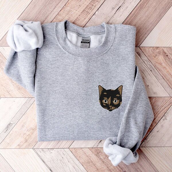 Sport Grey crewneck sweatshirt folded on a chevron wood floor with a tortoiseshell cat face, black with caramel patches, embroidered on the left chest