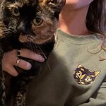 Customer holding her tortoiseshell cat while wearing a Military Green sweatshirt with the embroidered tortie cat face on the chest