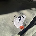 Close-up of the embroidered white ghost hugging a black cat with a jack-o'-lantern pail, bats and a crescent moon on a Military Green sweatshirt in sunlight