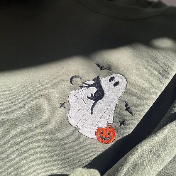Close-up of the embroidered white ghost hugging a black cat with a jack-o'-lantern pail, bats and a crescent moon on a Military Green sweatshirt in sunlight