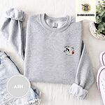 Ash crewneck sweatshirt folded with denim shorts, white sneakers and a potted plant, embroidered with a white ghost hugging a black cat and carrying a jack-o'-lantern pail under bats, a crescent moon and stars
